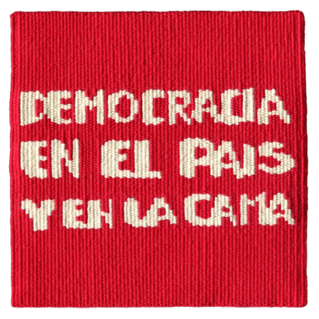 Imagen de un tapiz tejido a telar que reproduce la consigna feminista de la dictadura: “Democracia en el país y en la cama”. El texto está tejido con letras blancas sobre un fondo rojo.